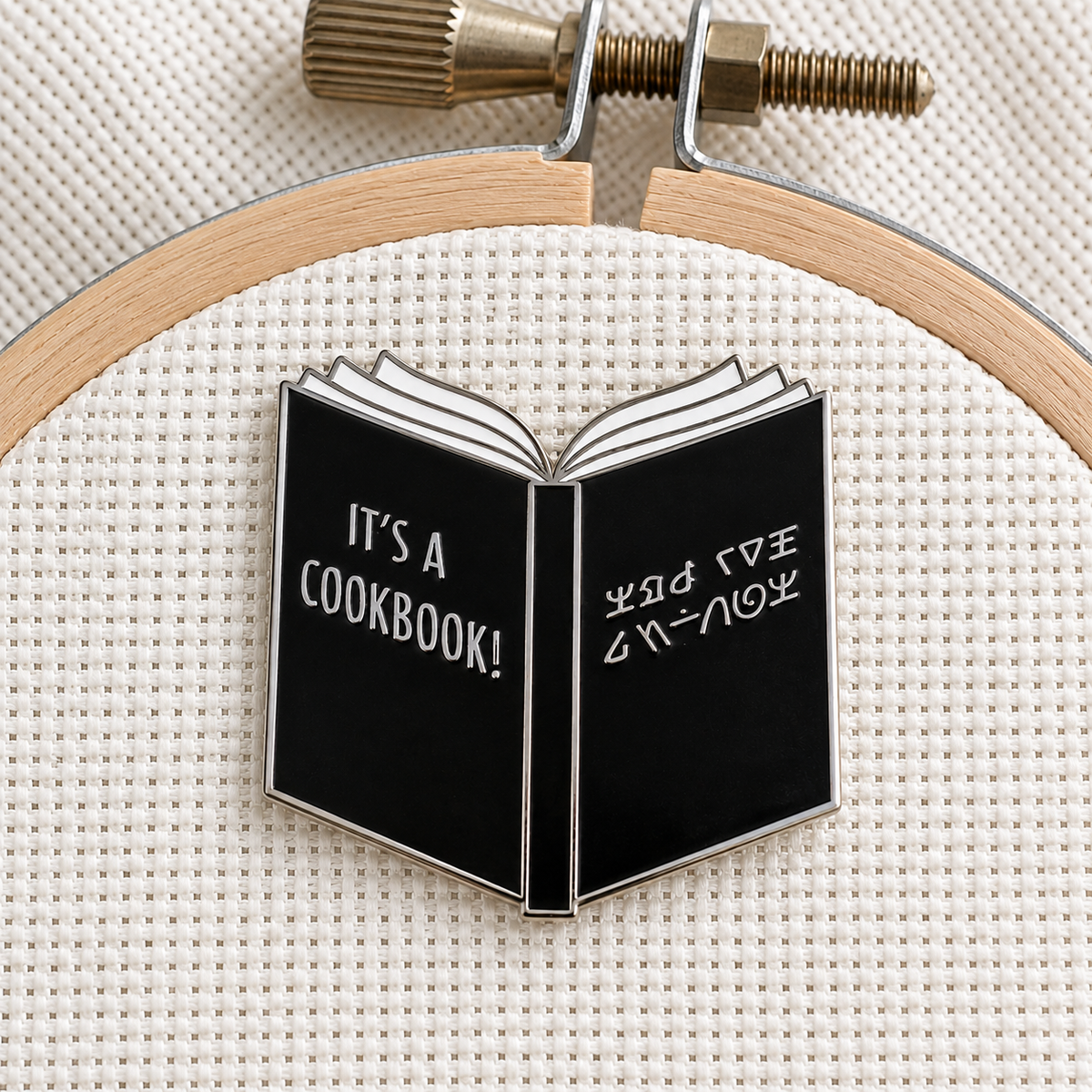 It's a Cookbook Needle Minder on an embroidery hoop, black open book with alien writing, silver finish