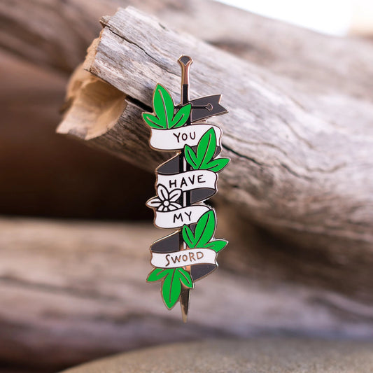 You Have My Sword pin propped against driftwood, outdoor sandy scene