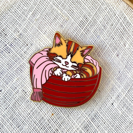 A Cat in a Basket pin — orange tabby kitten curled in a red striped basket, on white woven texture