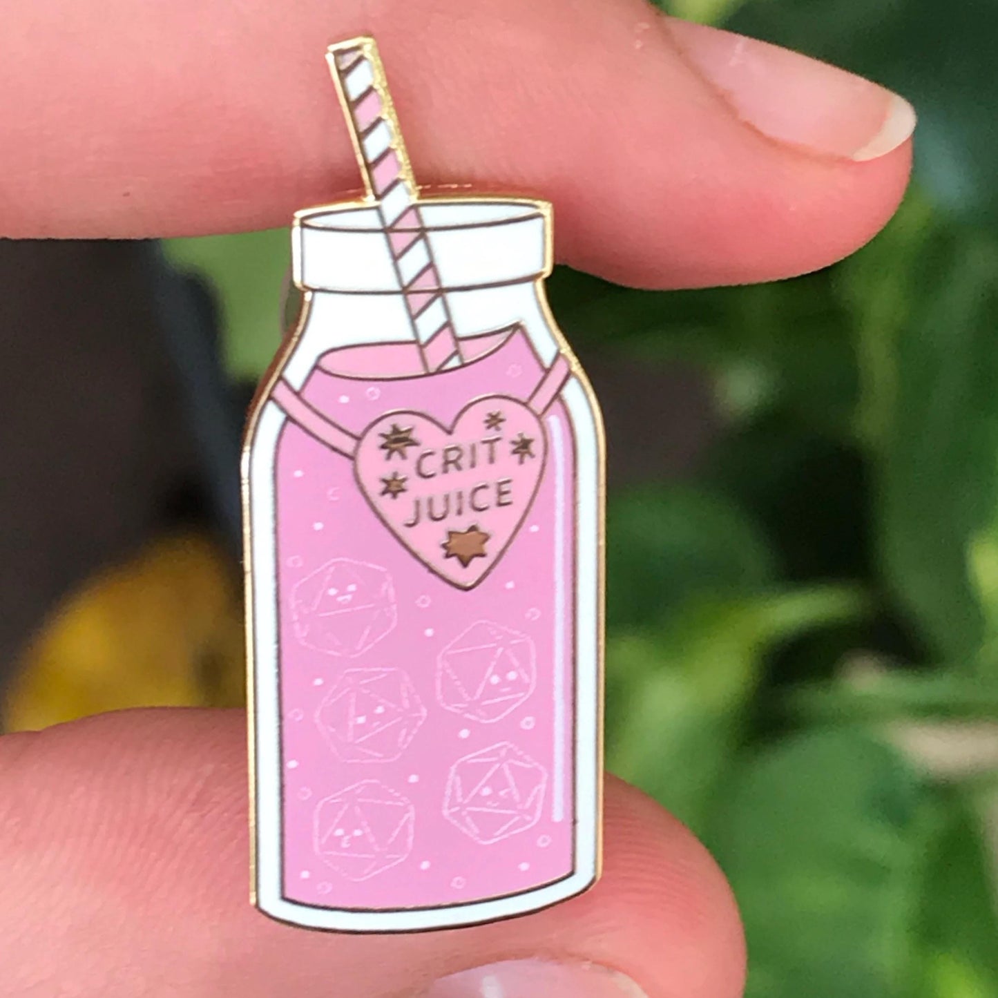 Crit Juice pin held in fingers — close-up showing pink juice with d20 outlines and the gold-trimmed heart 'CRIT JUICE' label.