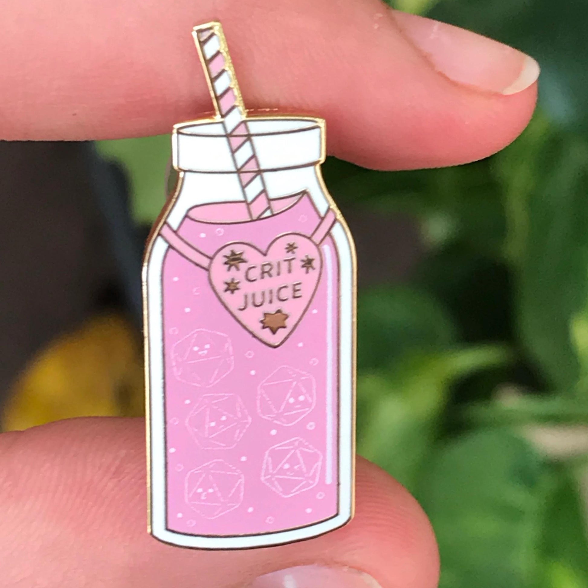 Crit Juice pin held in fingers — close-up showing pink juice with d20 outlines and the gold-trimmed heart 'CRIT JUICE' label.