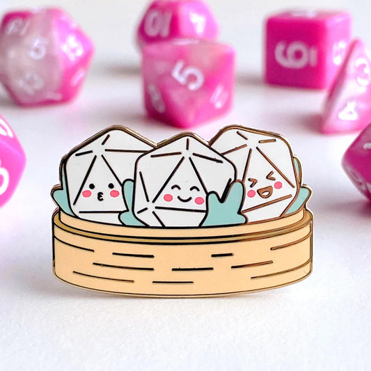 D20 Dim Sum pin — three white dumpling-shaped d20s with smiling faces sitting in a wooden bamboo steamer basket with teal leaves. Hard enamel pin photographed on a white surface with pink dice in the background. D&D kawaii pin.