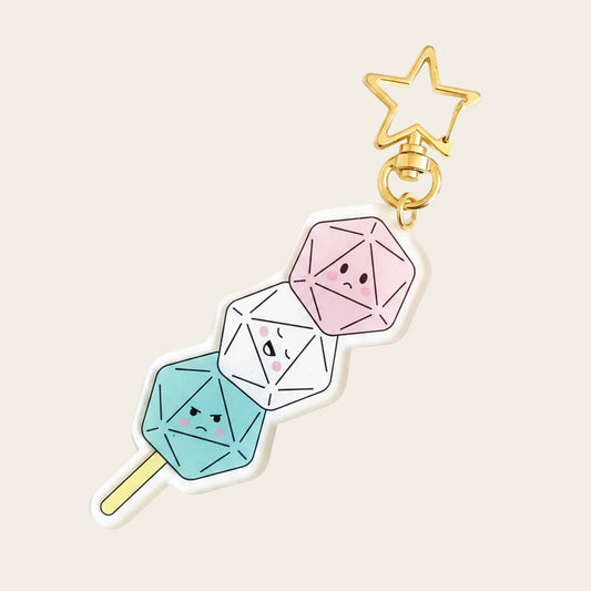 Dango Dice Charm — clear acrylic charm of three d20s on a dango skewer (pink, white, teal — happy, crying, grumpy faces), gold star keyring. Pink background with two real white d20s. D&D dice charm.