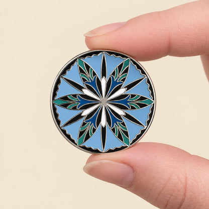 Sky blue elven cornflower enamel pin held between finger and thumb, silver nickel, about one inch across