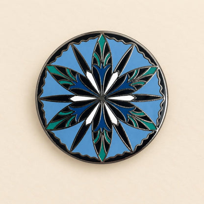 Round elven enamel pin, a twelve point cornflower in navy, teal and white on sky blue