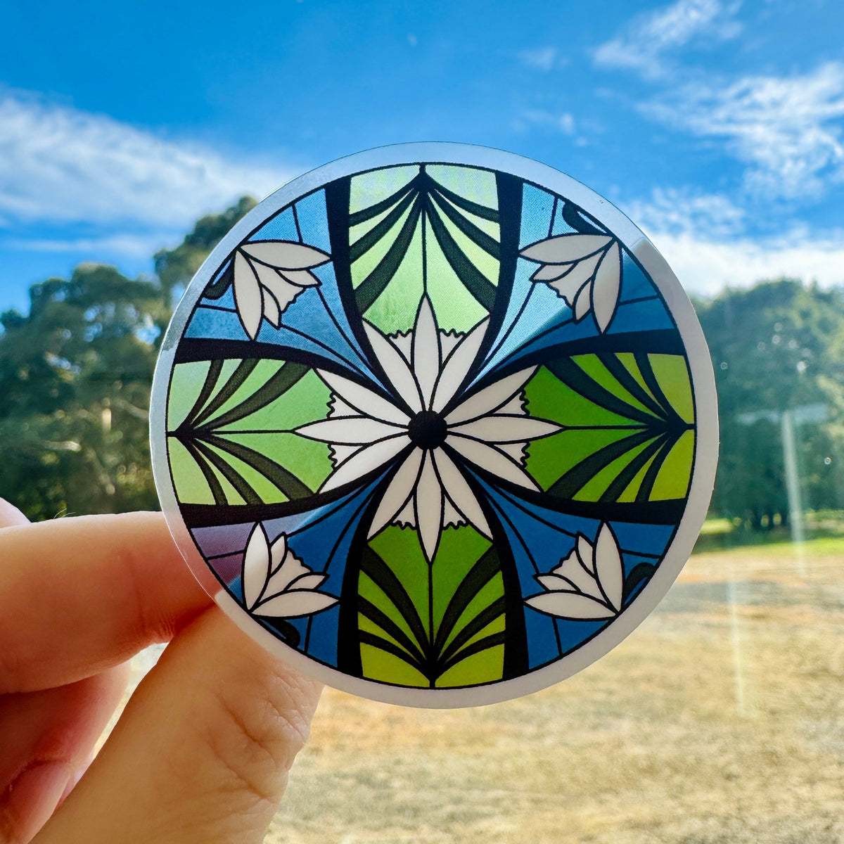 Elven Stained Glass Sticker, round clear sticker of green and blue stained glass with white flowers, held up to the sky, LOTR sticker