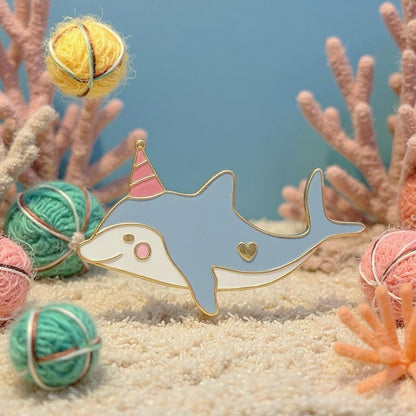 Party Dolphin enamel pin — gray dolphin with pink and red party hat, smiling, on grey wool background with felted yarn balls