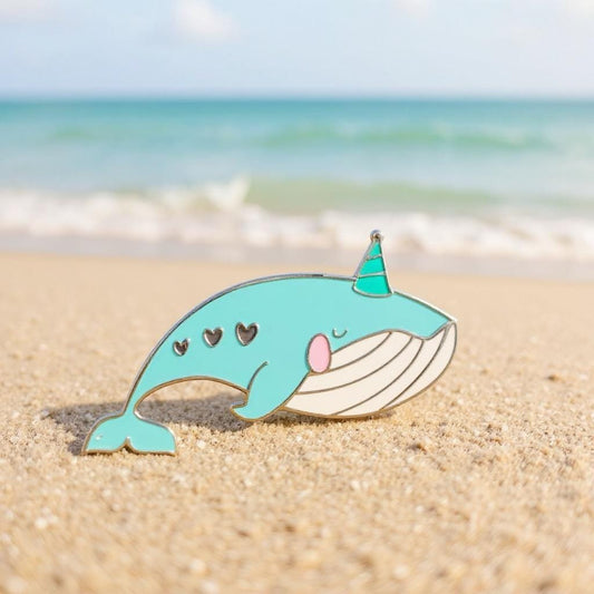 Party Whale enamel pin — teal whale with green party hat, hearts on cheek, sleepy eyes, on a beach with ocean in background