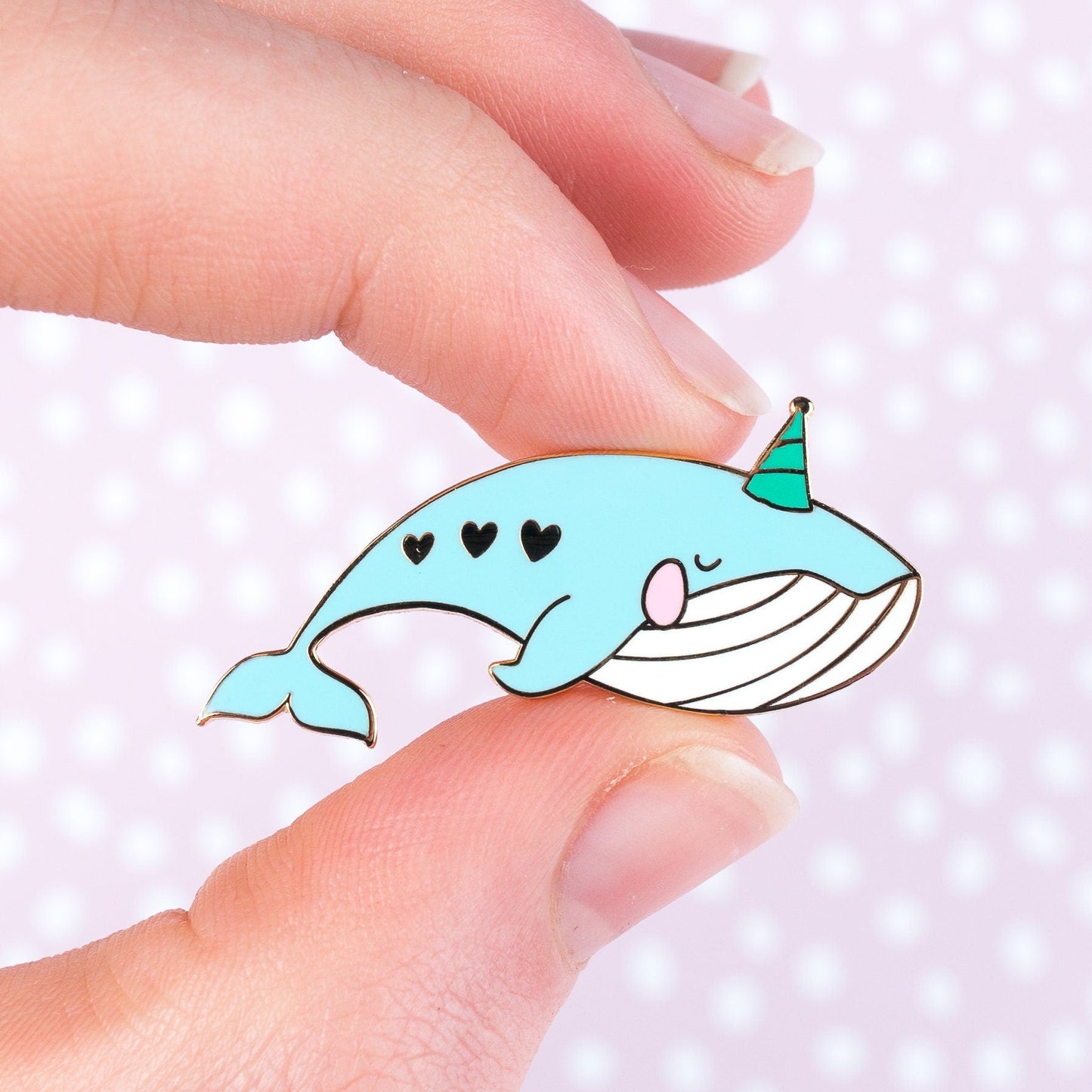 Party Whale enamel pin held in fingers against pink polka dot background