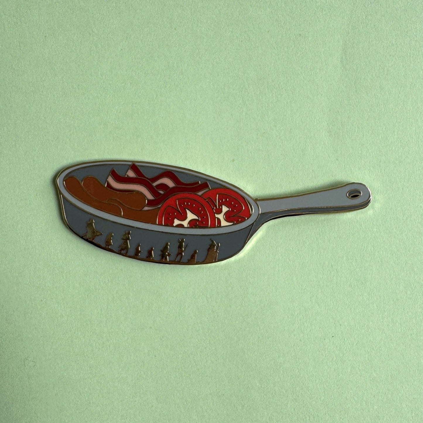 Sam's Saucepan pin on green - frying pan with bacon, sausages and tomato slices, Fellowship silhouettes on the side, LOTR enamel pin
