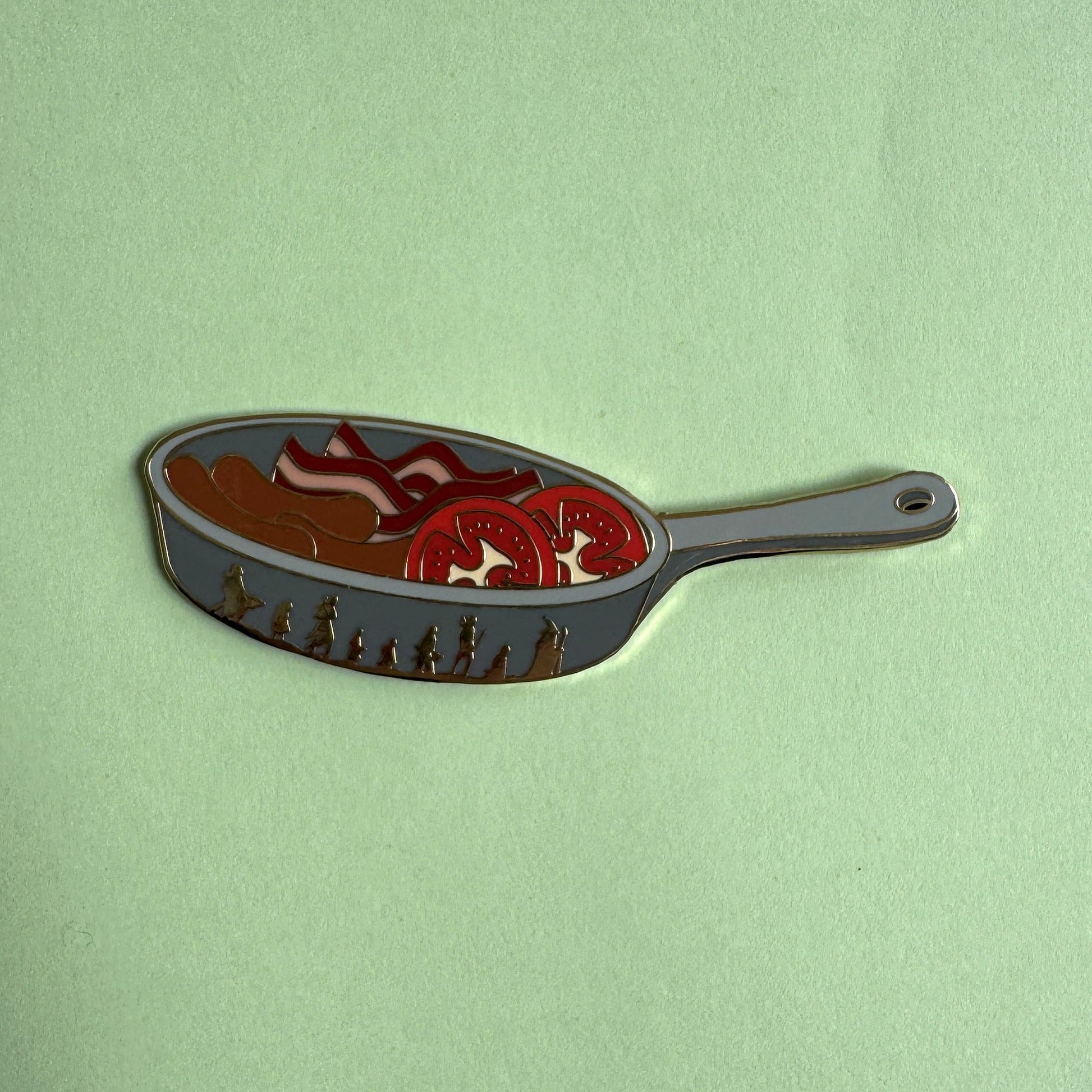 Sam's Saucepan pin on green - frying pan with bacon, sausages and tomato slices, Fellowship silhouettes on the side, LOTR enamel pin