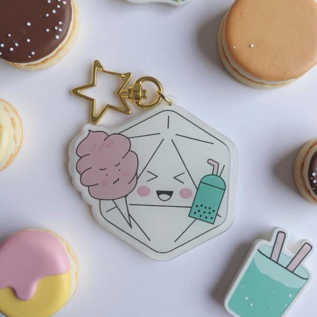 Yum Dice Charm — clear acrylic charm of a smiling d20 holding cotton candy and boba tea, gold star keyring. Lifestyle shot with iced cookies and macarons. D&D dice charm.